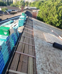 Roof under construction showing exposed wooden decking, structural support beams, and stacked piles of CertainTeed roofing shingles.