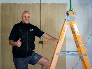 Smiling tradie in a Flow Master Plumbing and Gas branded work vest gives a thumbs up next to a fibreglass ladder on a job site.