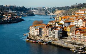 Aerial view of colorful historic buildings along the Douro River in Porto, Portugal, featuring traditional boats docked at the waterfront and an arched bridge in the background under a clear sky.