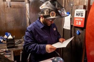 A welding competitor wearing protective gear reviews technical instructions inside a Project MFG welding booth during the 2026 National Welding League Championship.