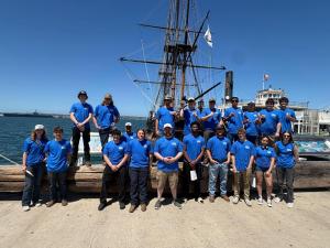 A group of students stand together in front of a ship wearing a blue project mfg t-shirt