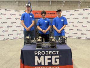 Three young men wearing blue Project MFG shirts stand behind a table displaying welded metal projects at a Project MFG event. Workshops for Warriors banners and an American flag are visible in the background.