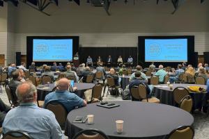 Wide shot of attendees seated at round tables during the Markets Matter Convening, with a speaker at a podium and panelists seated on stage between two event screens.