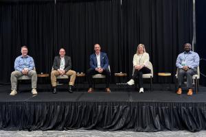 Five panelists seated on stage at the Markets Matter Convening, with microphones on small tables in front of a black curtain backdrop.