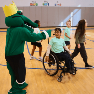 A green alligator masgot waves to a student in a wheelchair in the gymnasium.