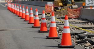 A long row of orange traffic safety cones lining an active road construction site with an excavator and road closed sign in the background.