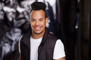 A smiling young man wearing a white shirt and black vest poses for a professional portrait against a softly blurred dark background. He has styled dark hair and is looking directly at the camera with a warm expression.