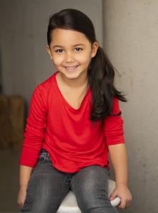 A smiling young girl wearing a bright red long-sleeve shirt and dark jeans sits on a stool during a professional portrait session. She has long dark hair tied to one side and is looking directly at the camera against a softly blurred neutral background.