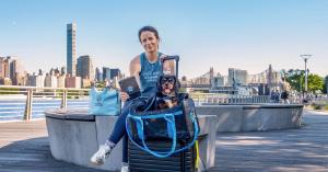 Lauren Anders Brown, CEO and founder of PadsPass, with her Cavalier King Charles Spaniel Peru in a pet carrier on a New York City waterfront, holding a tablet displaying the PadsPass app, with the Queensboro Bridge and Manhattan skyline in the background