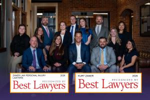Group photo of the Zaner Law Personal Injury Lawyers legal team in an office setting with a brick wall background. The bottom of the image features two official badges reading "Zaner Law Personal Injury Lawyers 2026 Recognized by Best Lawyers" and "Kurt Z