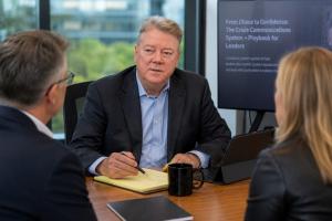 Crisis communications advisor Tim Brown meets with two executives in a corporate conference room while discussing crisis response strategy. A presentation screen displays information about the From Chaos to Confidence crisis communications system for lead