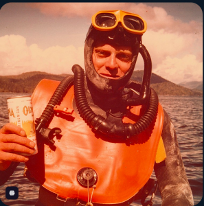 Gordon Esplin wearing an orange diving suit and gear on the waters of Vancouver Island, British Columbia, holding a drink after a dive.