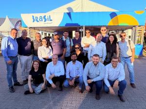 A group of professionals from the Brazilian delegation smiles as they pose in front of the Brazil stand at NAMPO Show in South Africa. In the background, the pavilion structure features the word “Brazil” and visual elements in blue, green and yellow, refl
