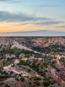 Sunset photo of Palo Duro Canyon at MERUS Adventure Park, with a line of off-road vehicles and Jeeps with headlights on descending a winding dirt switchback trail through the red and tan canyon walls, surrounded by green juniper and mesquite, beneath a pa