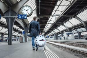 Person walking through a railway station carrying a large model of the Official Swiss Railways Station Clock beneath a station clock.