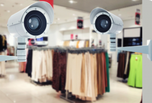 Two white security surveillance cameras in the foreground monitoring a blurred retail clothing store layout with clothing racks, illustrating defensive security spending on local commerce. Image created by mytsv.com.