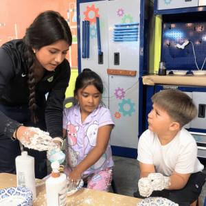 A woman guides two children in a playful science experiment with foam and lotion. The room has colorful lockers and a creative atmosphere.