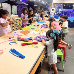 Children engaged in arts and crafts, working at a large table with colorful materials in a bright, creative workshop space.