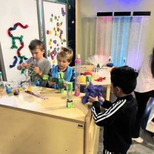 Children engaged in hands-on STEM activities with colorful tubes, gears, and light-up sensory panels in an interactive learning space.