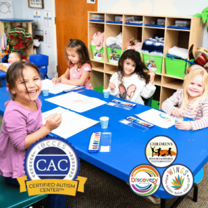 Four children sit around a blue table using watercolors to paint in a colorful and organized classroom.