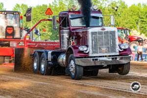 Maroon Peterbilt semi truck pulling a competition sled on a dirt track.
