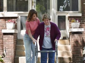 An adult daughter helping her elderly mother down the front steps of a brick house using a walker, highlighting senior living transitions and downsizing in Chicagoland.