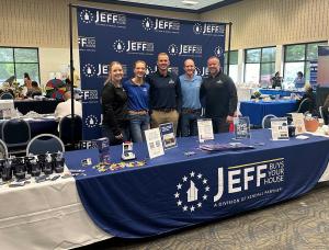Five smiling team members from Jeff Buys Your House standing behind a blue branded table and backdrop at a local community senior expo in Chicagoland.