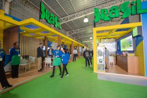 An exhibition space from Brazil at Exponor appears in a large trade fair pavilion, with booths in green, yellow, and blue and large suspended signs reading “Brasil.” Around 12 people are walking around and talking in the area, some wearing badges and casu