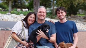 Puppy Raiser family holding their puppy for the first time, a man, woman and teenage son sitting on a bench with the grass and GDA|TLC campus in background.