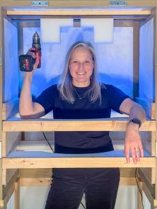 Heather Haldeman, Senior Client Advisor at Atelier 4, smiling and holding a power drill while standing inside a partially built wooden art shipping crate lined with blue protective foam.