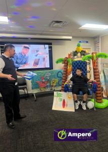 Ampere Electric Owner Shane Viggiani standing next to Make-A-Wish child Luis seated, during an indoor presentation.