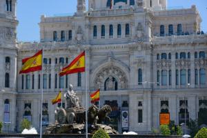 The red and yellow national flag of Spain waves on a flagpole next to the ornate stone tower of a historic classical building under a clear blue sky.