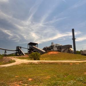 Wide view of an industrial forest products facility with conveyors, processing structures and a tall stack under a blue sky, with grassy fields in the foreground.