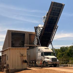 White semi-truck unloading wood fiber at a biomass facility using a raised hydraulic truck tipper beside a processing building.
