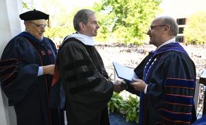 James G. Basker shakes hands with Gettysburg College President Bob Iuliano while accepting an honorary degree during Gettysburg College's commencement ceremony. Three individuals in academic regalia are pictured on stage.