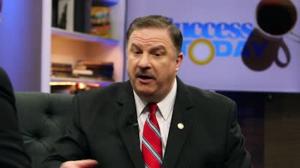 Richard Tyler is sitting on a chair in a dark blue suit with a red tie with blue stripes. His gold Commitment to Excellence® logo lapel pin is visible. In the background is a Success Today TV monitor.