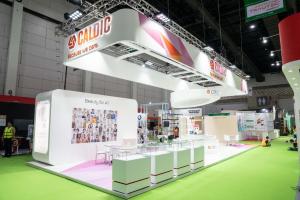 CALDIC because we care exhibition booth at in-cosmetic Asia featuring large diversity photo wall, minimalist white design, glass display cases, and modern truss structure showcasing inclusive beauty solutions
