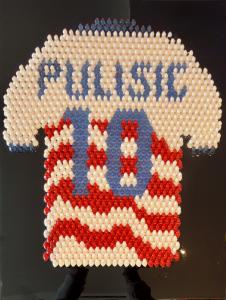 Sculpture of a USA soccer jersey spelling Pulisic 10 formed from rows of red white and blue tiny resin ducks on a black background
