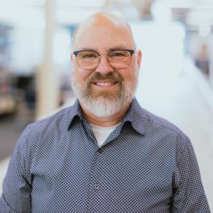 Headshot of a smiling person wearing glasses, and a beard and mustache