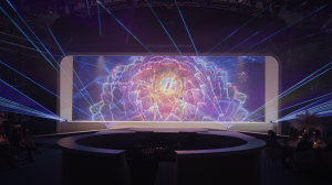 Two dancers in white perform in front of a wide LED screen displaying a vibrant pink and purple bloom, with blue lasers fanning outward across the venue.