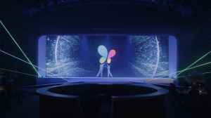 Two dancers in white reach upward together in front of an LED screen showing a multicolored butterfly emerging from a digital particle field, with green lasers crossing the stage.