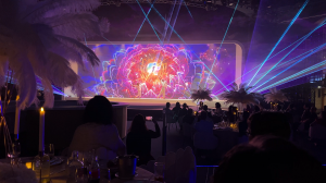 View from the back of the venue showing the stage and a vivid floral visual on the LED screen, with audience members silhouetted at candlelit tables among ostrich-feather centerpieces and lasers crossing the room overhead.