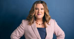 Whitney Pennington, Founder of Pennington Law Firm, PLLC, poses for a professional headshot wearing a light pink blazer and blue blouse, standing confidently with her hands on her hips against a blue background.