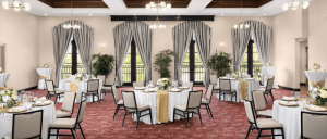 Elegant banquet room with round tables, white tablecloths, gold runners, upholstered chairs, large windows, and potted plants.