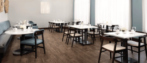 Modern empty restaurant dining area with wooden chairs, white tabletops, large windows, and soft natural lighting.