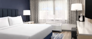 Bright hotel room with a large bed, bedside lamp, floor lamp, window with curtains, and modern furniture.