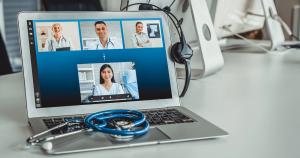 photo of a large computer screen sitting on a table, on the screen are large squares with photos of health care professionals online in each screen signifying telehealth