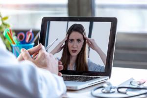 photo of a health care professional at their desk at a lap top, on the screen of the computer you can clearly see a patient, they are conducting a telehealth patient session