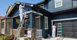 Two JLM Painting crew members painting the exterior of a dark blue two-story home in Petaluma, California, with ladders, paint buckets and drop cloths visible.
