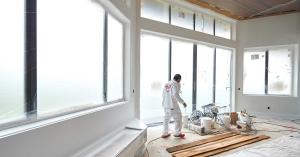 A professional painter wearing white overalls walks past an airless paint sprayer and Benjamin Moore paint buckets inside a bright luxury home in Petaluma. The large floor-to-ceiling windows are completely masked off with plastic sheeting and tape to prot